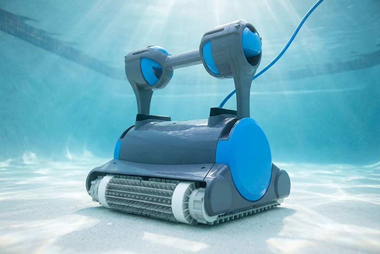 A Dolphin Premier robotic cleaner on the floor of a pool, a slate-gray body with a round bright blue side cover, gray and white brush rollers across the front, and a tall gray handle with a blue float at each end and a blue cable rising behind it