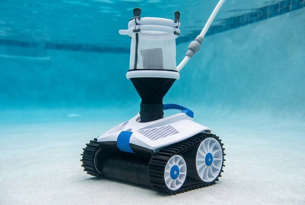 A pressure-side pool cleaner on the floor of a pool, a white body with blue trim and a blue handle on black rubber tank tracks with white spoked wheels, a see-through white mesh debris canister on a black cone on top, and a white hose with a gray swivel rising from it
