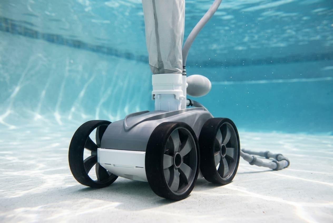 A four-wheeled pressure-side pool cleaner on the floor of a light plaster pool, a gray top over a white front and wide black tires, with a gray mesh debris bag and a round float rising from its top and a gray sweep hose trailing behind it