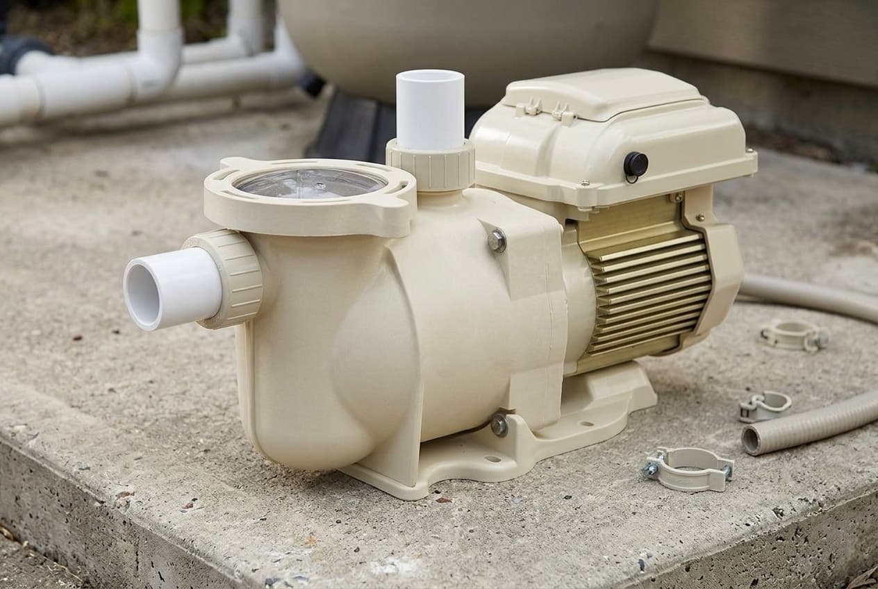 An almond pool pump on a concrete equipment pad with union fittings at both ports and a clear strainer lid on top, hose clamps and flexible conduit laid beside it mid-install