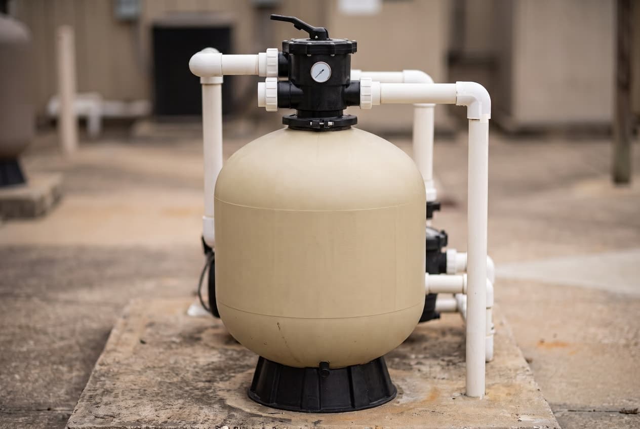 A rounded almond sand filter tank on a black flared foot on a concrete pad, a black multiport valve on top with a pressure gauge on its front and white PVC pipes running from its three ports