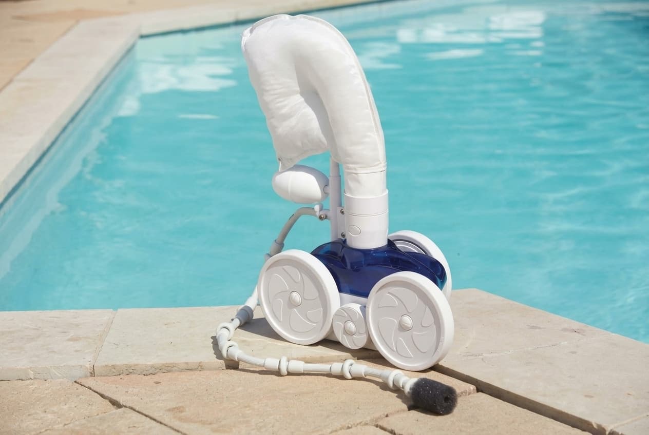 A blue-bodied pressure-side pool cleaner on stone coping at the water's edge, its white debris bag standing up from the mast, three white wheels, and the sweep hose trailing behind with its wear rings and dark scrubber tip