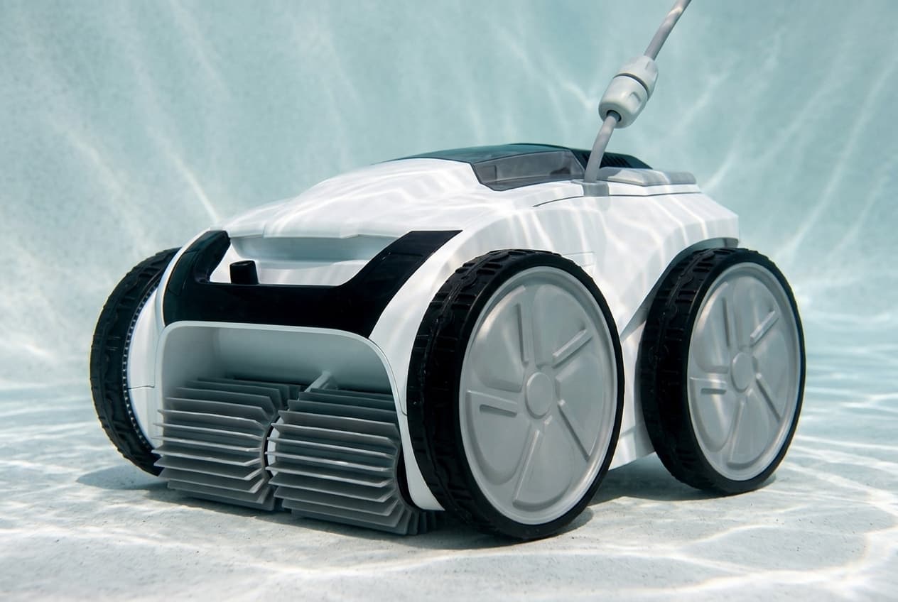 A white Polaris VRX iQ+ robotic cleaner on the floor of a pool, black trim across its front above twin gray bladed brushes, four gray wheels with black tires, a smoked lid on top and a gray cable rising through a pale gray swivel