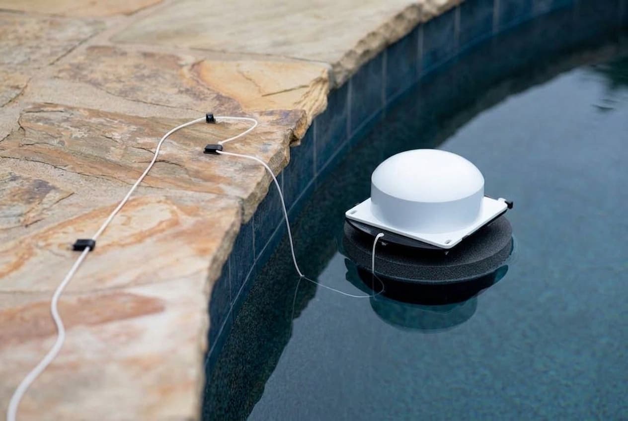 A white dome-topped floating pool alarm on a square white base and a round black foam float, drifting in dark water beside flagstone coping, a thin white tether running from it up onto the stone